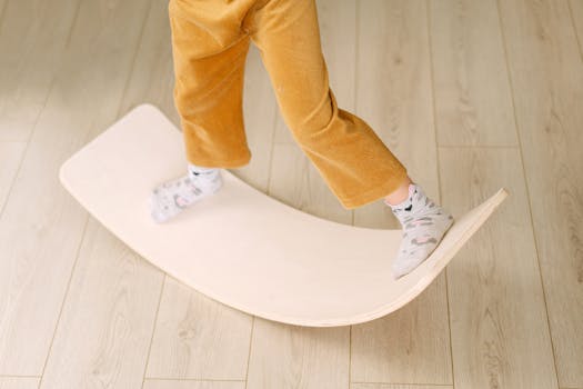 A child wearing yellow pants balances on a wooden balance board indoors.